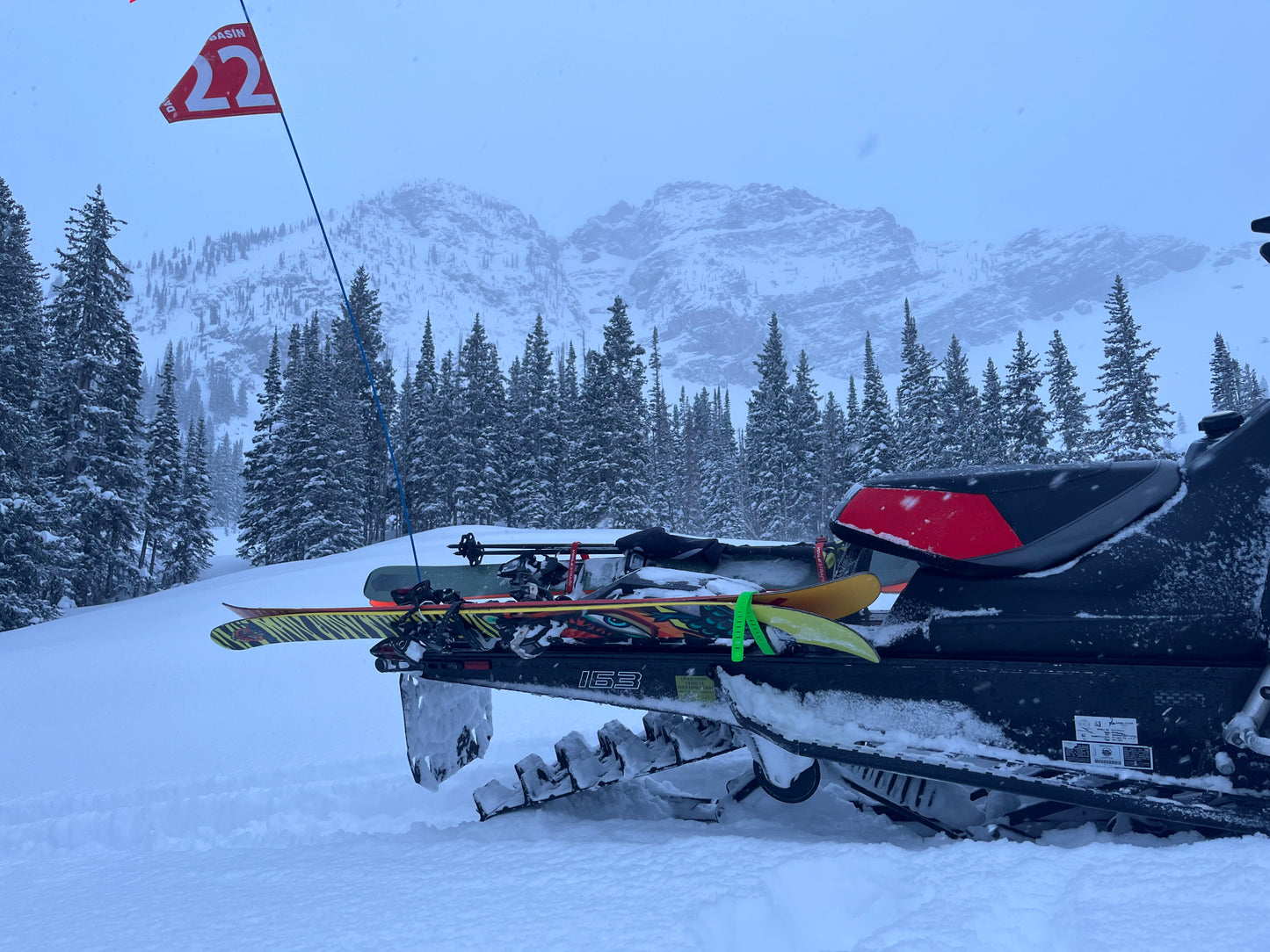 A close-up of a snowmobile equipped with a backcountry ski rack system, securely holding a pair of skis along the side. The skis are fastened with sturdy brackets and straps, positioned to avoid interference with the tunnel bag, which sits neatly behind the seat. Snow-covered tracks and a powdery trail in the background highlight the rugged, off-trail setting.