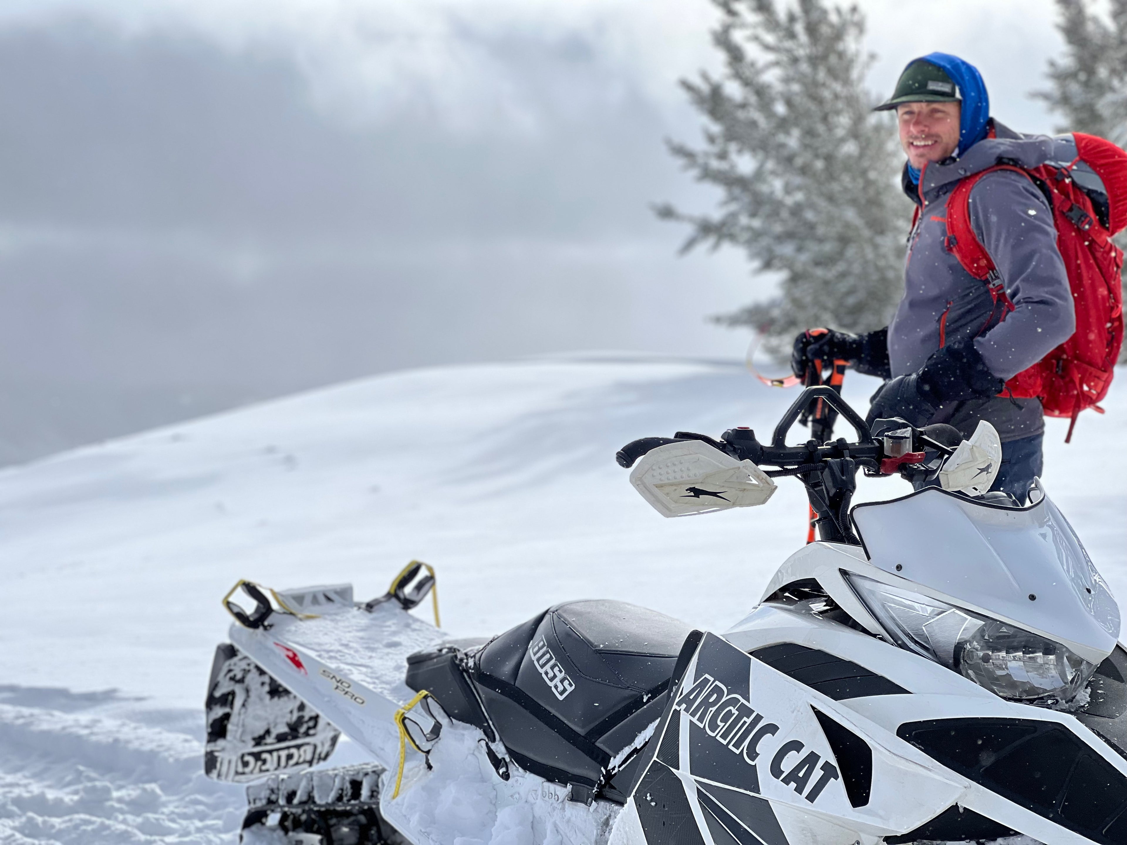 A backcountry skier stands next to a white Arctic Cat snowmobile in a snow-covered alpine setting, wearing a red backpack and holding ski poles. The snowmobile is equipped with a specialized ski rack system, allowing access to remote terrain that was previously inaccessible. Snow-covered hills and trees in the background emphasize the deep winter environment and the freedom this system provides for exploring untouched backcountry areas.
