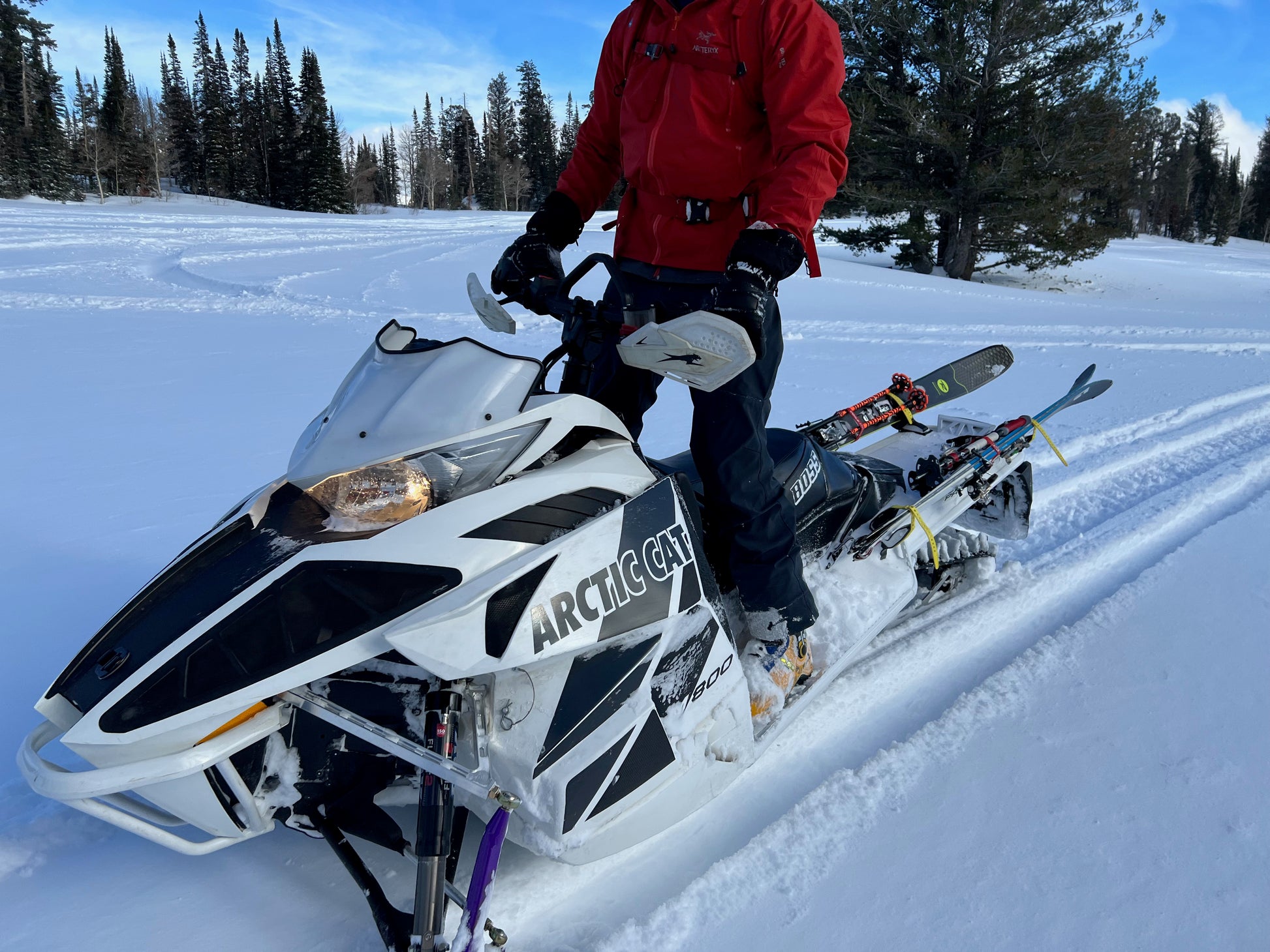 A backcountry skier stands next to a white Arctic Cat snowmobile in a snow-covered alpine setting, wearing a red backpack and holding ski poles. The snowmobile is equipped with a specialized ski rack system, allowing access to remote terrain that was previously inaccessible. Snow-covered hills and trees in the background emphasize the deep winter environment and the freedom this system provides for exploring untouched backcountry areas.