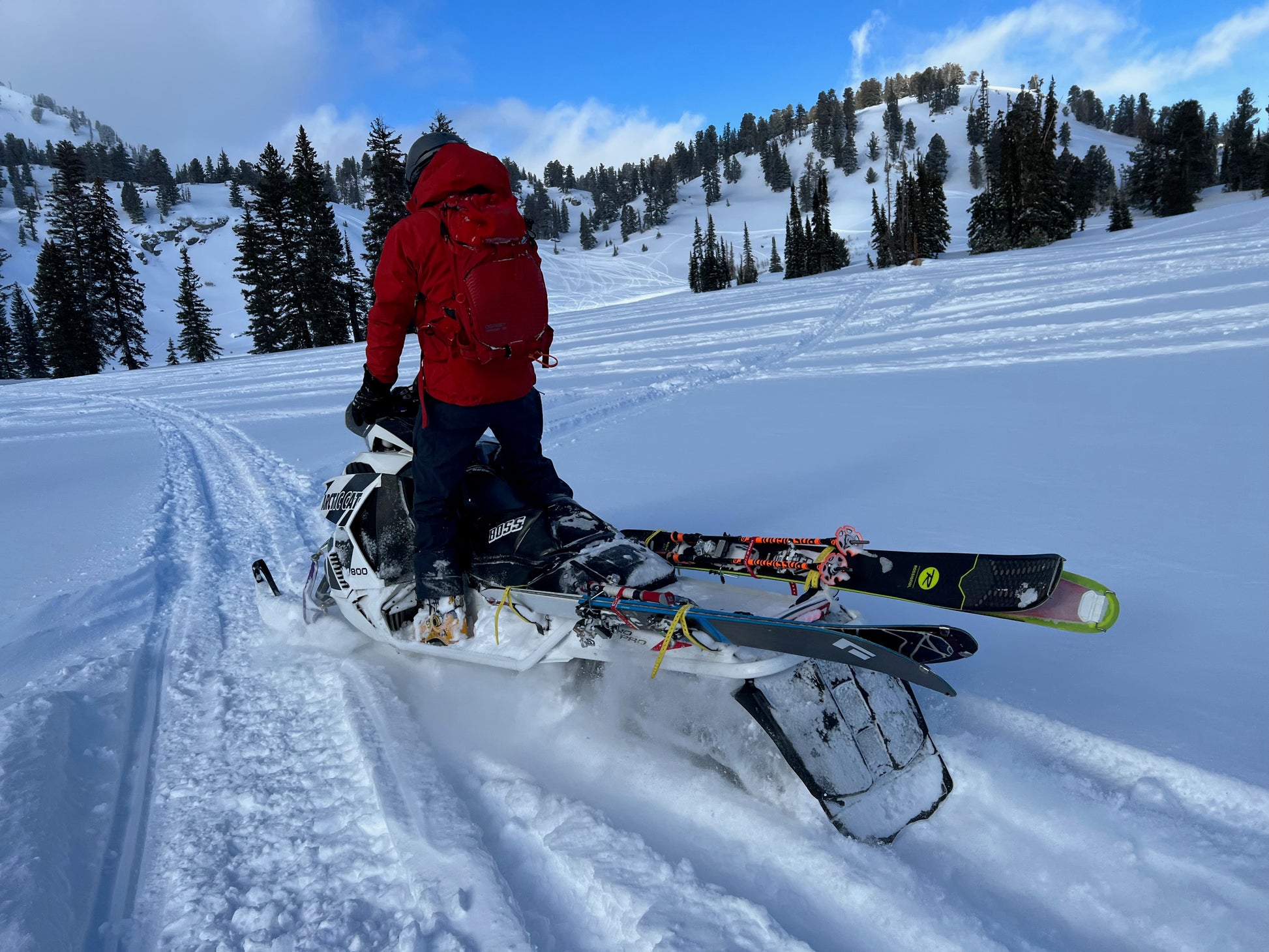 A backcountry skier stands next to a white Arctic Cat snowmobile in a snow-covered alpine setting, wearing a red backpack and holding ski poles. The snowmobile is equipped with a specialized ski rack system, allowing access to remote terrain that was previously inaccessible. Snow-covered hills and trees in the background emphasize the deep winter environment and the freedom this system provides for exploring untouched backcountry areas.
