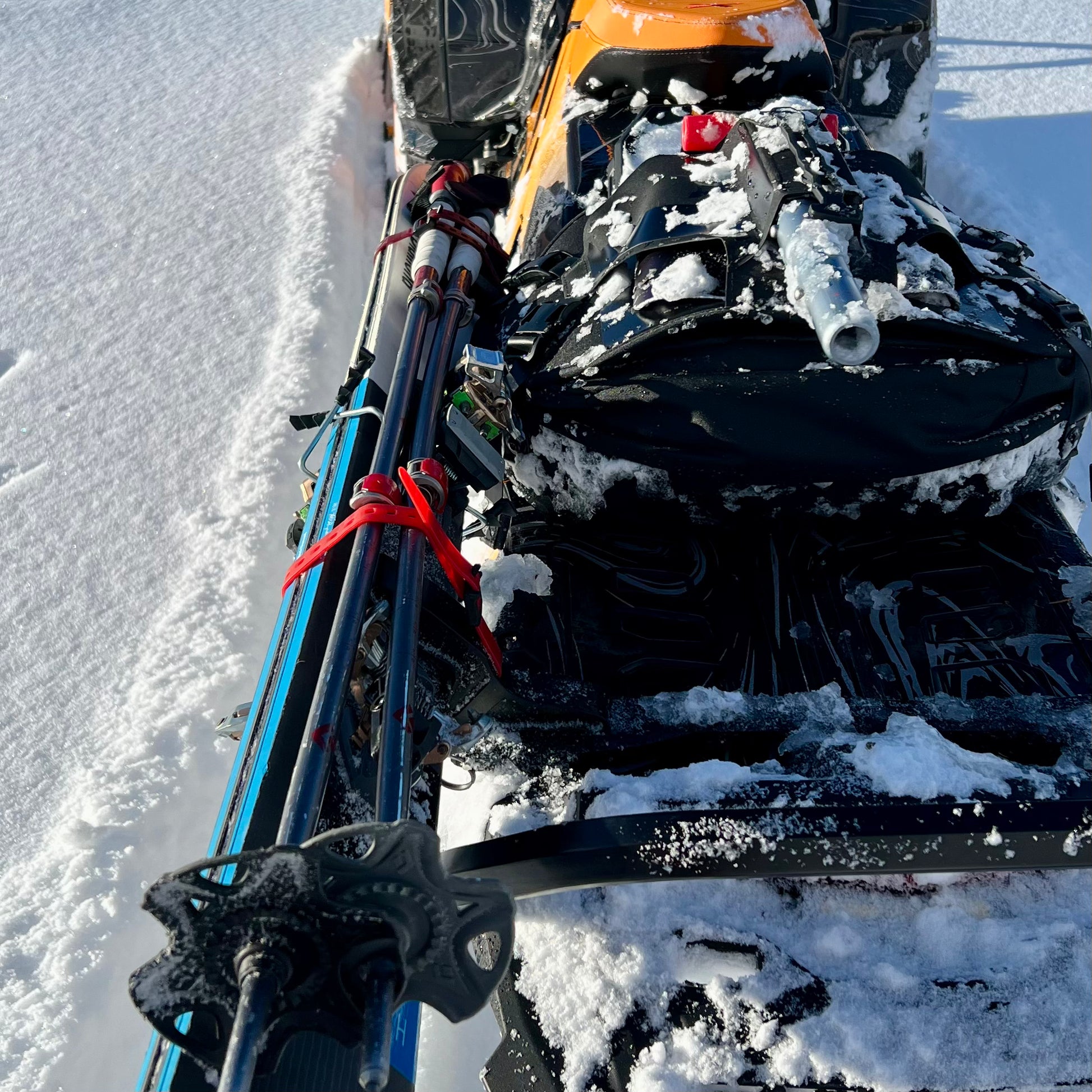 A close-up of a snowmobile equipped with a backcountry ski rack system, securely holding a pair of skis along the side. The skis are fastened with sturdy brackets and straps, positioned to avoid interference with the tunnel bag, which sits neatly behind the seat. Snow-covered tracks and a powdery trail in the background highlight the rugged, off-trail setting.