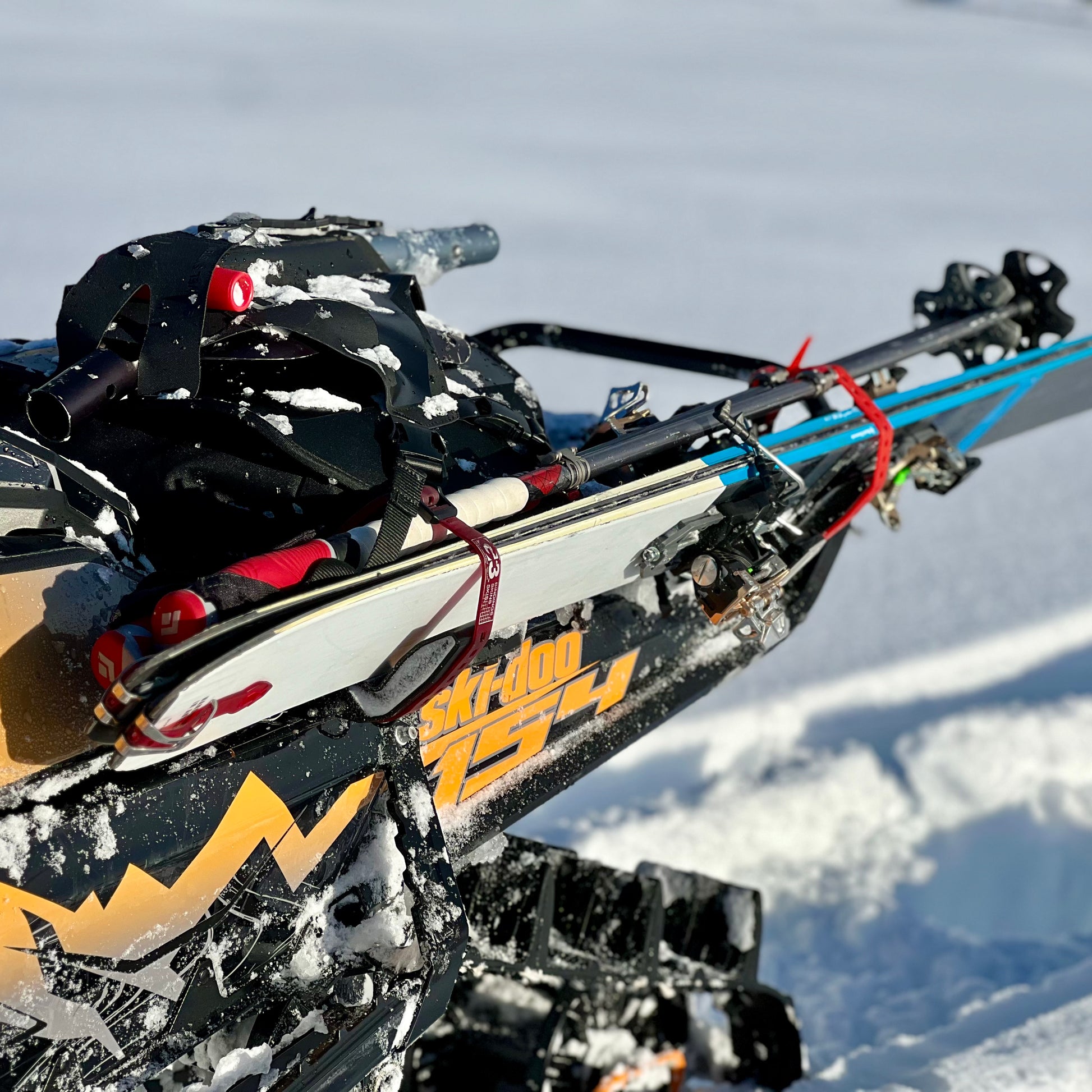 A close-up of a snowmobile equipped with a backcountry ski rack system, securely holding a pair of skis along the side. The skis are fastened with sturdy brackets and straps, positioned to avoid interference with the tunnel bag, which sits neatly behind the seat. Snow-covered tracks and a powdery trail in the background highlight the rugged, off-trail setting.