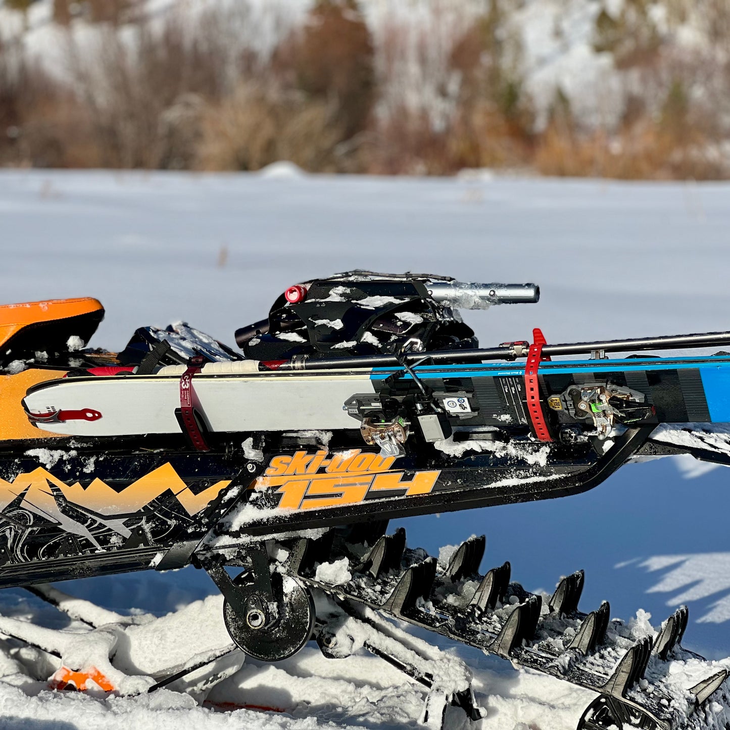 A close-up of a snowmobile equipped with a specialized rack system designed for carrying skis in backcountry conditions. The skis are securely fastened to the side of the snowmobile using sturdy straps and brackets, ensuring stability during off-trail travel. The snowmobile's track and suspension are visible, partially covered in snow, with a scenic winter landscape in the background.