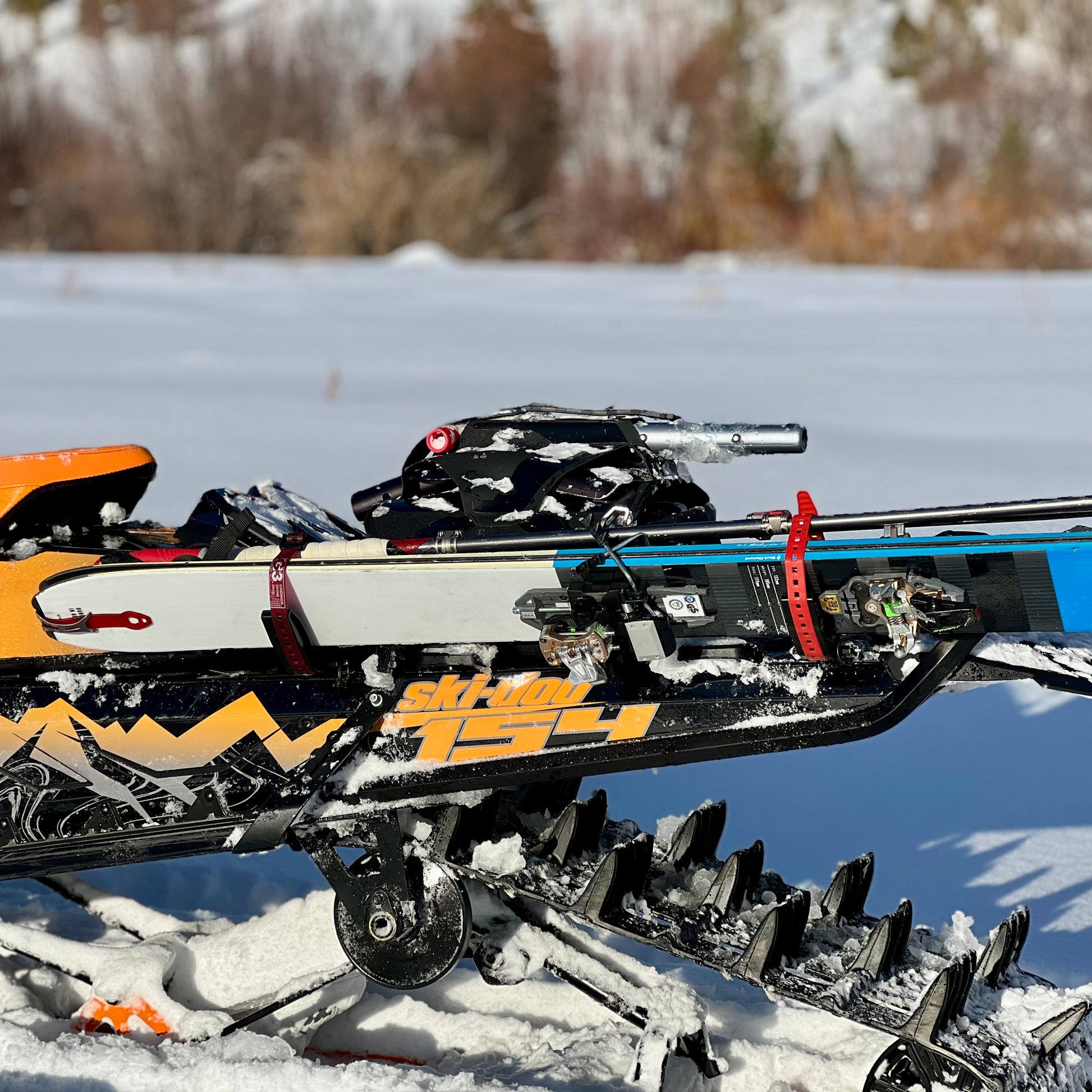 A close-up of a snowmobile equipped with a specialized rack system designed for carrying skis in backcountry conditions. The skis are securely fastened to the side of the snowmobile using sturdy straps and brackets, ensuring stability during off-trail travel. The snowmobile's track and suspension are visible, partially covered in snow, with a scenic winter landscape in the background.