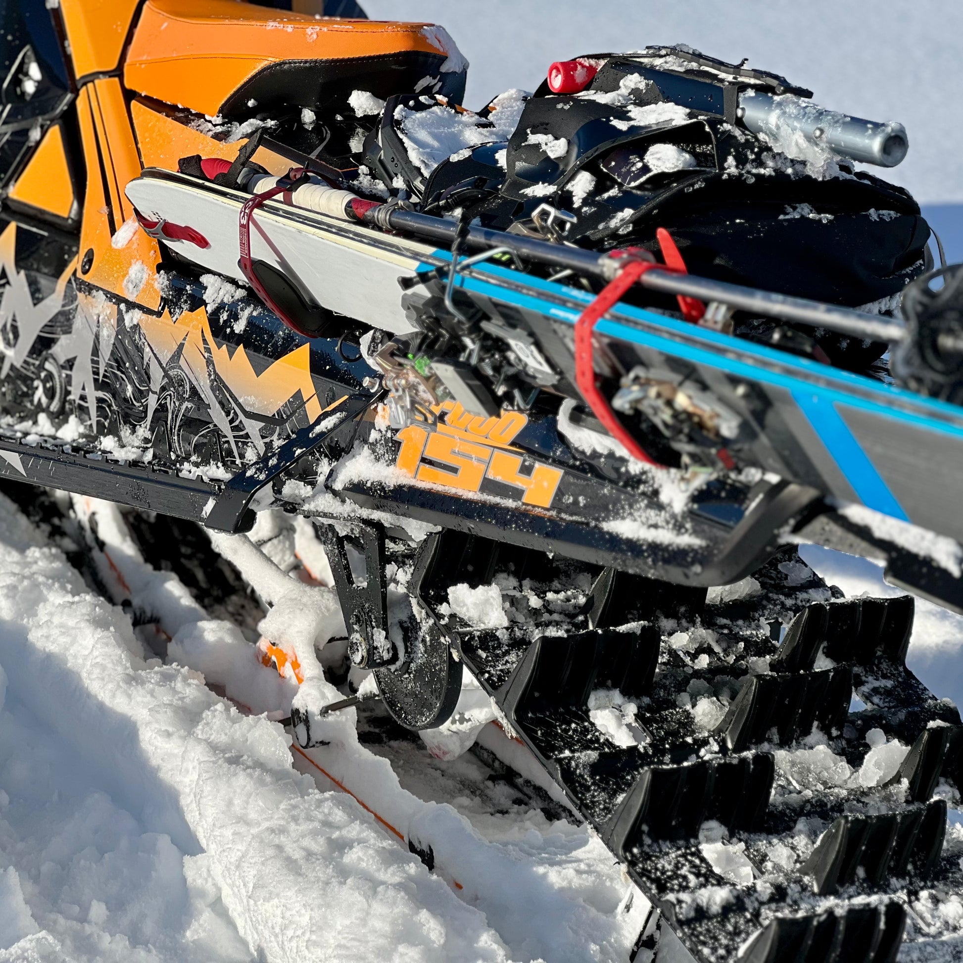 A close-up of a snowmobile equipped with a backcountry ski rack system, securely holding a pair of skis along the side. The skis are fastened with sturdy brackets and straps, positioned to avoid interference with the tunnel bag, which sits neatly behind the seat. Snow-covered tracks and a powdery trail in the background highlight the rugged, off-trail setting.