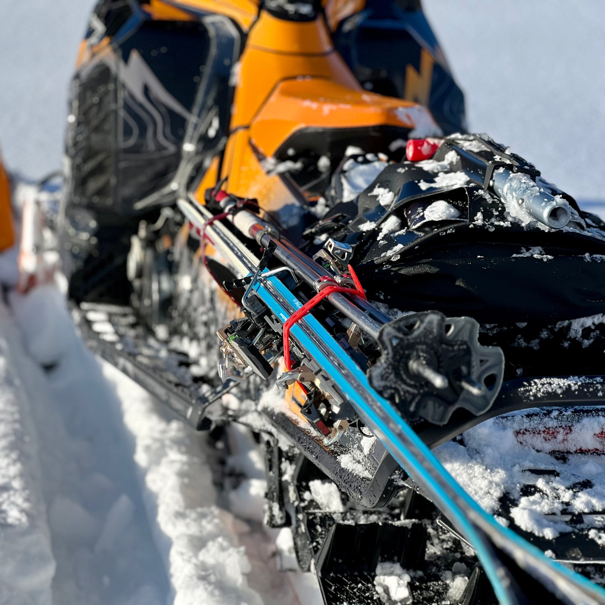 A close-up of a snowmobile equipped with a backcountry ski rack system, securely holding a pair of skis along the side. The skis are fastened with sturdy brackets and straps, positioned to avoid interference with the tunnel bag, which sits neatly behind the seat. Snow-covered tracks and a powdery trail in the background highlight the rugged, off-trail setting.