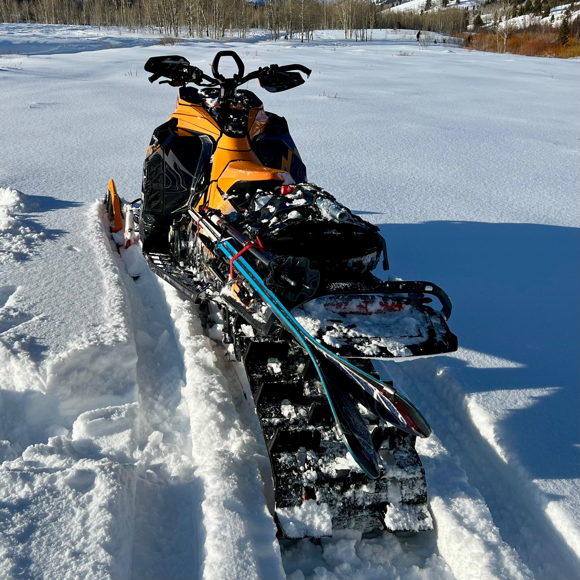 A close-up of a snowmobile equipped with a backcountry ski rack system, securely holding a pair of skis along the side. The skis are fastened with sturdy brackets and straps, positioned to avoid interference with the tunnel bag, which sits neatly behind the seat. Snow-covered tracks and a powdery trail in the background highlight the rugged, off-trail setting.
